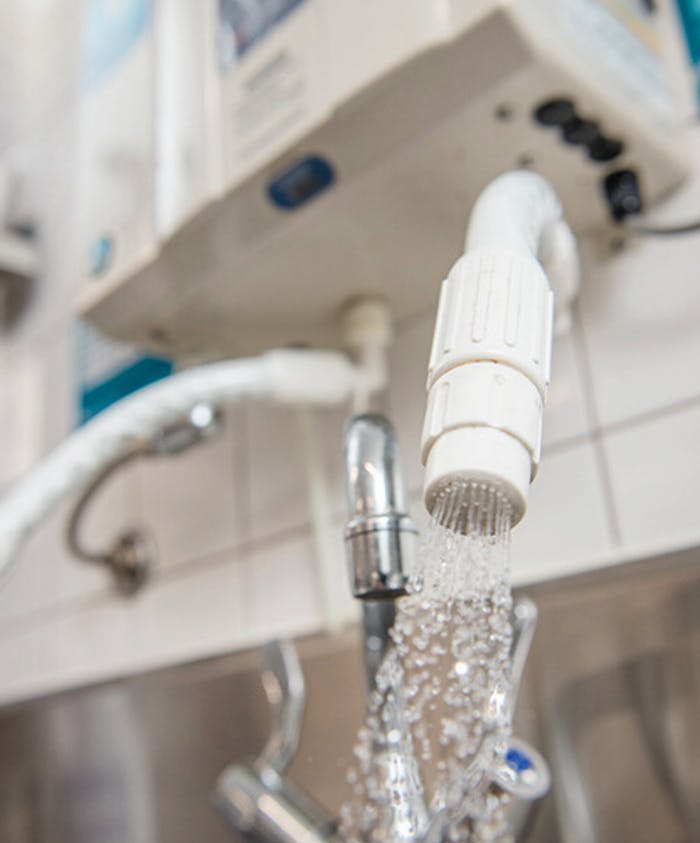 A white, cylindrical water filter emits a steady stream of water in a stainless-steel kitchen setting. The Te Pae brand appears on the device mounted above, indicating its purpose and usage.